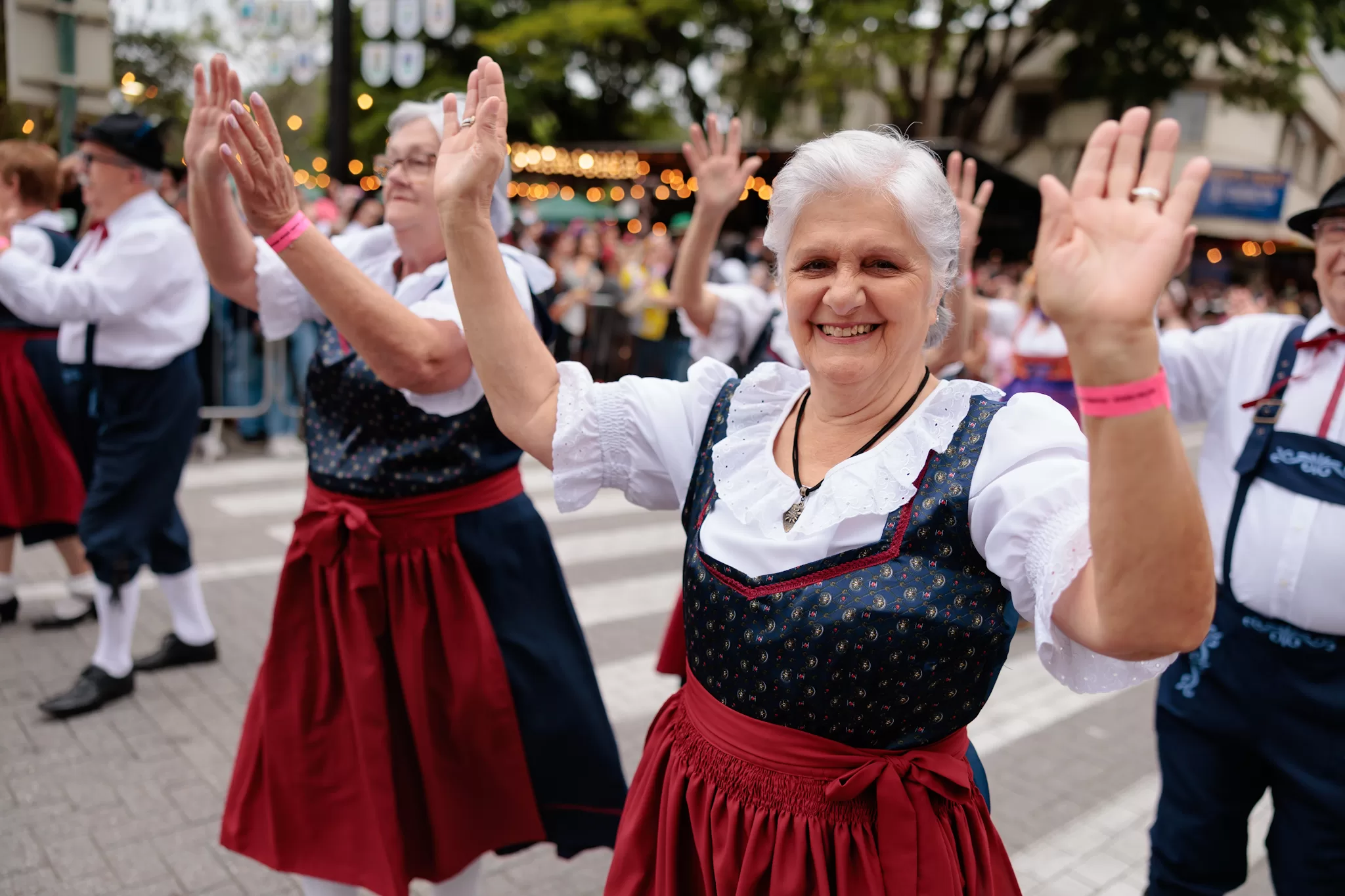 Foto no desfile da Oktoberfest. Na imagem aparece duas idosas, a primeira olha para a fota com as mãos para cima e sorri. Elas estão vestidas com trajes típicos alemães